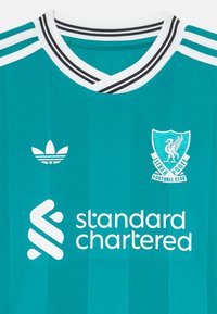 Maglia da calcio color turchese con strisce bianche e scollatura a V. Presenta il logo Adidas, il distintivo del Liverpool FC e il marchio "Standard Chartered".