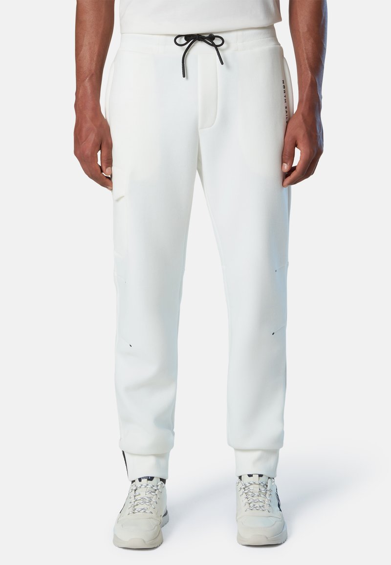 Pantalones sudorosos blancos fabricados en una mezcla de algodón, con cintura ajustable, bolsillos laterales y puños acanalados. Llevados con zapatillas de color claro.