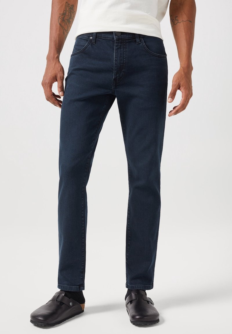 Man i mörkblå slim fit-jeans, svarta strumpor, och svarta slip-on skor med spännen, stående på en ljus bakgrund.