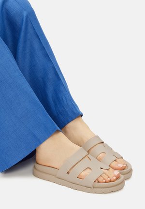 Beżowe sandały na płaskiej podeszwie z wyciętym wzorem, wykonane z gładkiego materiału, z grubą podeszwą i szerokimi paskami. Noszone z niebieskimi spodniami o fakturze.