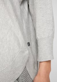 Detailní záběr na šedou pletenou svetr s rukávem a lemem, na boku malá černá textilní štítka, oblečeno s texturovanými šedými kalhotami a viditelným předloktím.