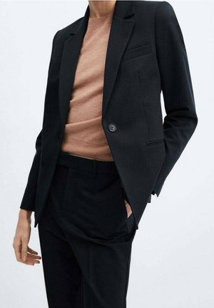 Personne portant un blazer noir et un pantalon avec un pull beige, une main dans la poche, sur un fond clair uni.