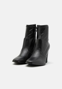 Botas de cuero negro hasta el tobillo con punta afilada, tacón alto y detalle de cremallera. Textura suave con acentos de costura a lo largo de las costuras.