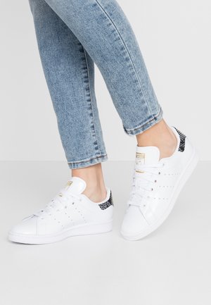 Osoba w jasnoniebieskich dżinsach i białych trampkach z czarnym, fakturowanym detalem na obcasie, na neutralnym tle.