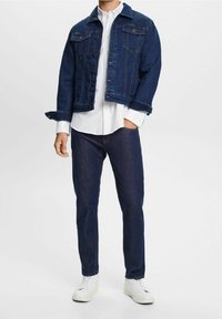Dunkelblaue Jeansjacke mit Knopfverschlüssen, zwei Brusttaschen und einem leicht verkürzten Saum. Kombiniert mit einem weißen Hemd und dunklen Hosen.