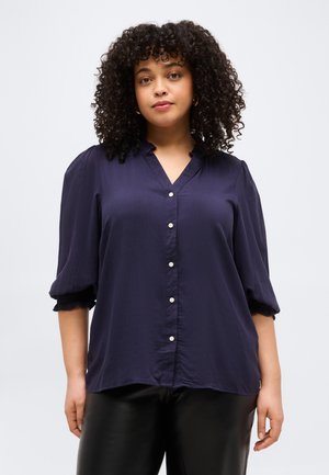 Frau mit lockigem Haar, die eine marineblaue, geknöpfte Bluse mit dreiviertellangen Ärmeln und schwarze Hose vor neutralem Hintergrund trägt.