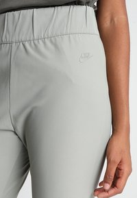 Světlé šedé sportovní kalhoty s hladkou texturou, elastickým pasem a embosovaným logem Nike na stehně. Přiléhavý střih a minimální detaily.