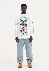 Langärmliges weißes Hemd mit buntem grafischem Design, das ein Kreuz und Text zeigt. Kombiniert mit hellblauen Jeans und braunen Stiefeln.