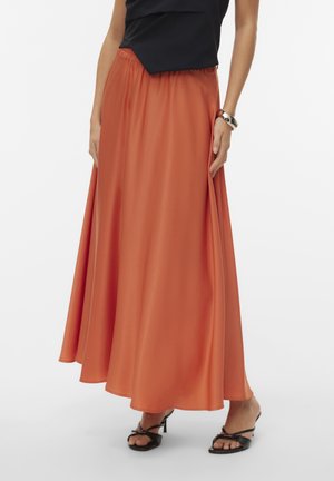 Vrouw draagt een lange vloeiende rok in verbrande oranje kleur, een zwarte mouwloze top, zwarte open pumps en een grote zilveren armband om haar pols.