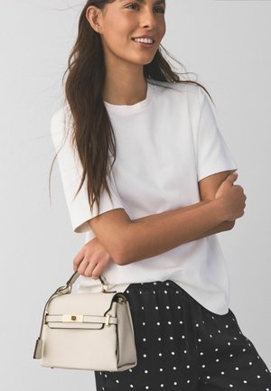 Bēšs rokassomiņa ar zelta detaļām, strukturētu dizainu un augšējo rokturi. Valkāts ar baltu kreklu un melnām biksēm ar baltām punktiņām.