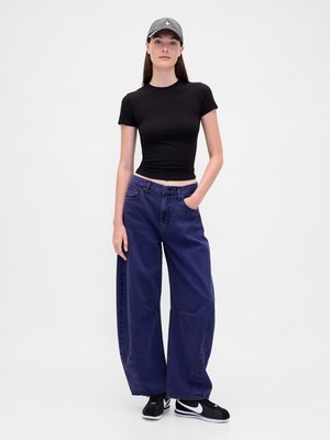 Mlada žena nosi crni uski crop majicu, široke tamnoplave traperice, crne Nike tenisice i sive kape na jednobojnoj pozadini.