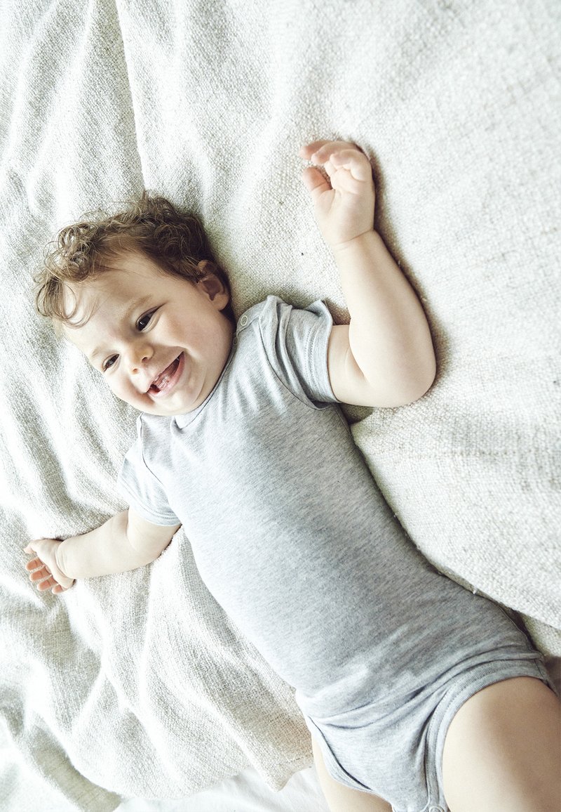 Grå bomulds-body på en baby, der ligger på et tekstureret, lyst tæppe. Babyen har krøllet hår og armene strakt ud, hvilket får den til at se glad ud.