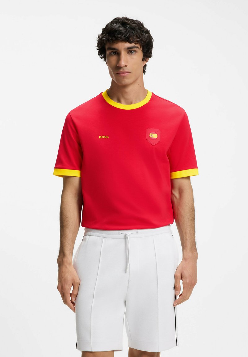 Jeune homme portant un t-shirt rouge avec bordure jaune et emblème, associé à un short blanc, debout devant un fond blanc uni.