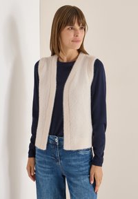 Ljusbeige ärmlös väst gjord av fluffigt material, med V-ringning och utan knäppningar, bärs över en mörk marinblå långärmad topp.