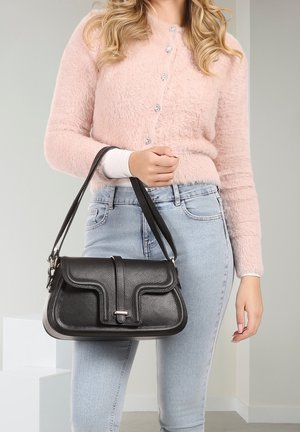 Vrouw in een lichtroze wollige trui en lichtblauwe spijkerbroek, die een zwarte leren schoudertas met dubbele riemen vasthoudt.
