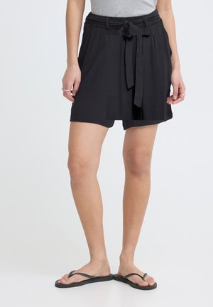 Kvinde iført sorte, højtaljede shorts med et bundet bælte og sorte flip-flops, stående mod en ensfarvet hvid baggrund.