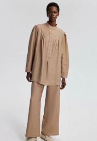 Beige, textured outfit featuring a long-sleeve, button-up top with pleats and loose-fitting pants. Includes subtle buttons and a high collar.