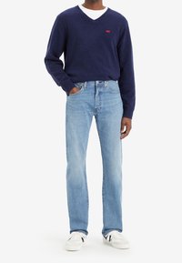 Suéter azul marino con cuello en V, jeans de pierna recta color azul claro, camiseta blanca debajo, zapatillas blancas con detalles en negro. Detalle de logo rojo en el suéter.