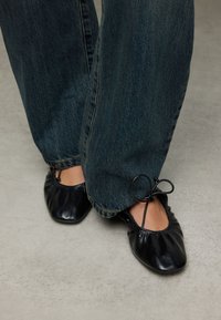 Zwarte leren ballerina's met geplooide details en een decoratieve strik. Gecombineerd met donkerblauwe wijde jeans. Gladde textuur, afgeronde neus.