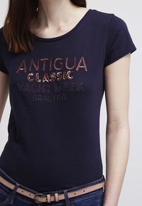 Námořnická modrá bavlněná tričko s kulatým výstřihem, krátkými rukávy a vyšitým nápisem "ANTIGUA CLASSIC YACHT WEEK GAASSTRA" v bronzových a metalických nitích.