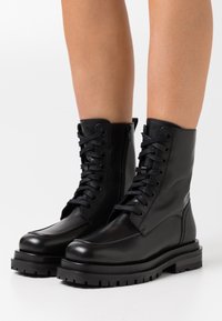 Bottines en cuir noir avec lacets et fermeture éclair latérale. Semelle en caoutchouc épaisse avec finition texturée. Tige lisse avec des détails de couture minimaux.