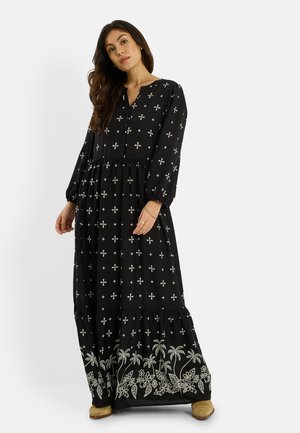 Femme portant une robe maxi noire à manches longues avec des motifs blancs en croix et floraux, debout devant un fond clair uni.