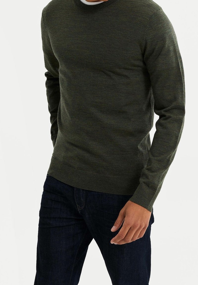 Homme portant un pull vert foncé à col rond et un jean bleu foncé, debout devant un fond clair uni.