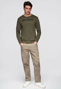 Oliwkowa bluza z napisem "OMBRE" na piersi, zestawiona z jasnobeżowymi spodniami i białymi tenisówkami, stojąca na jednolitym tle.