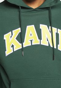 Grön hoodie med känguruficka, utrustad med vit och gul broderad text "KANI" över bröstet. Dragbanden har metallagletter.