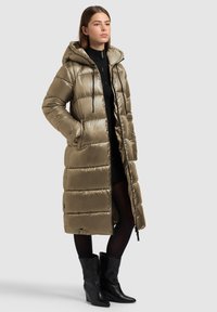 Lange, gewatteerde jas in een gedempte olijfgroene kleur, met een capuchon, horizontale doorgestikte lijnen en steekzakken aan de zijkant. Draagt over een zwart gebreide jurk en panty's.