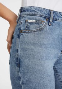 Light blue denim jeans with a high waist, featuring subtle fading, yellow stitching, and a Guess Jeans label on the back pocket.