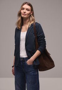 Femme blonde portant un blazer bleu marine, un haut blanc, un jean bleu, avec un grand sac à bandoulière marron, debout avec une main dans la poche, fond neutre.