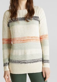 Pull en tricot avec des rayures horizontales en crème, vert, corail et noir. Encolure et poignets côtelés épais. Matériau doux et texturé.