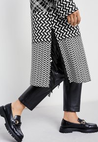 Svart och vitt mönstrad kappa med fransdetaljer, kombinerad med glänsande svarta läderbyxor och loafers med metallaccent.