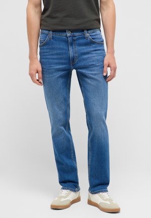 Mann trägt mittelblaue Jeans mit geradem Bein und weiße Sneakers mit beigen Akzenten, steht vor einem schlichten hellen Hintergrund.