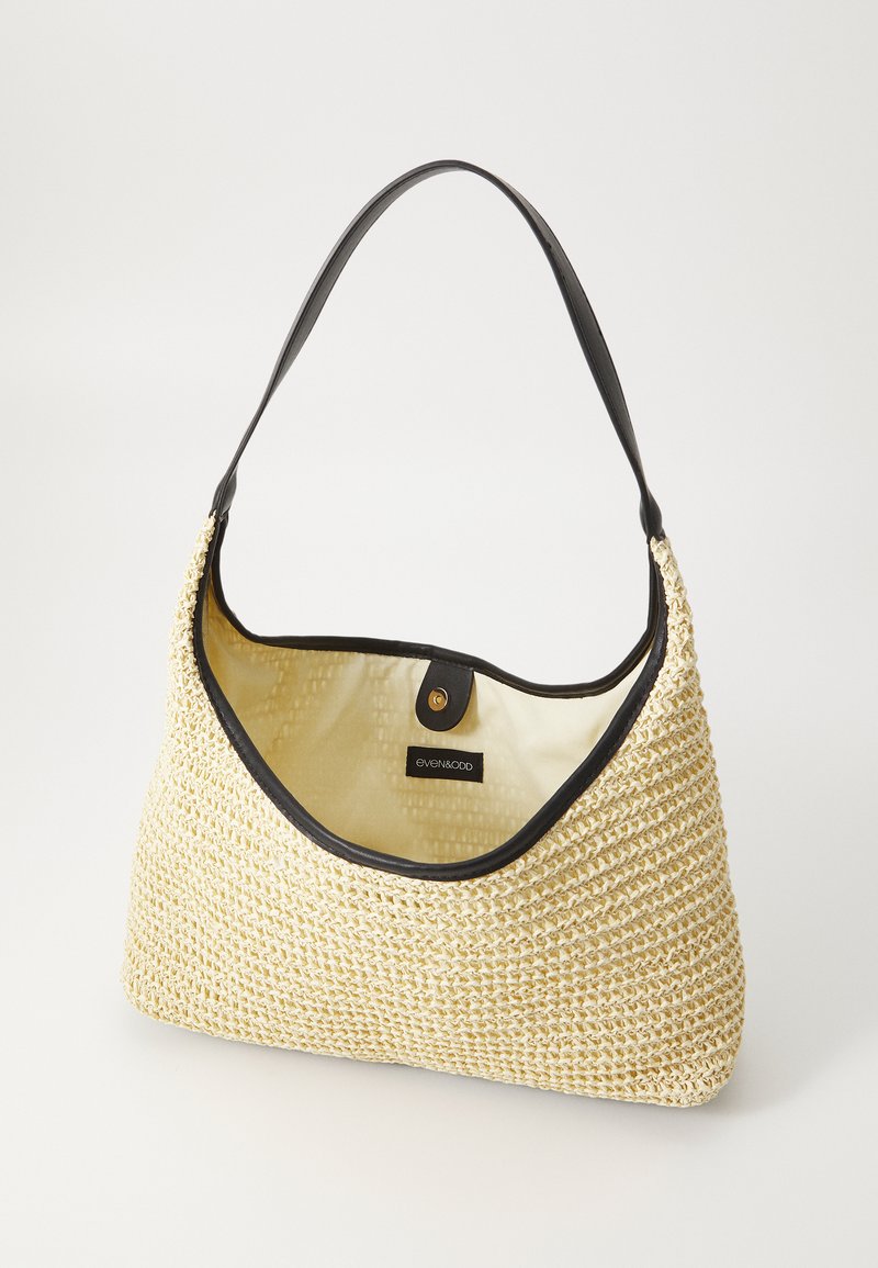 Bolso de hombro de paja trenzada con un ribete de cuero negro, presenta una textura suave, forma curva y un cierre a presión. El interior es beige.