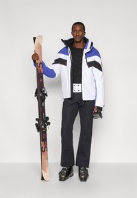 Lyžiarsky outfit s vodeodolnou bundou v bielej, modrej a čiernej farbe, čierne nohavice s textúrovanými detailmi a lyžiarskym vybavením, vrátane lyží a topánok.