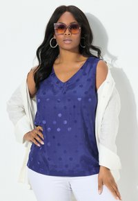 Top sin mangas de color azul marino con escote en V y un patrón de lunares, combinado con una camisa blanca ligera abierta, presentada por un modelo.