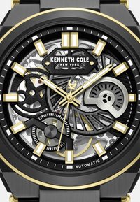 Automatyczny zegarek Kenneth Cole z czarną i złotą kopertą, otwartą tarczą ukazującą skomplikowane zębatki oraz wyraźnymi znacznikami godzin w kolorze złotym i srebrnym.
