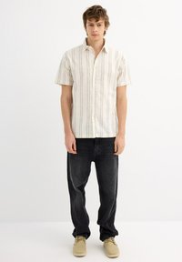 Camisa de botones de manga corta color crema con finas rayas verticales, combinada con jeans negros holgados y zapatos beige. Diseño sencillo y corte relajado.