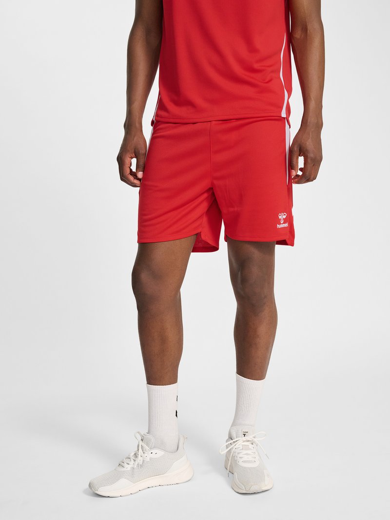 Uomo con pantaloncini sportivi rossi, maglietta rossa, calzini bianchi e scarpe da ginnastica bianche, in piedi su uno sfondo chiaro uniforme.
