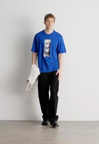 Μπλε κοντομάνικο μπλουζάκι με γραφικό σχέδιο, συνδυασμένο με μαύρα cargo παντελόνια. Μια λευκή πλεκτή δημιουργία είναι ριγμένη σε ένα χέρι.