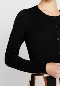 Svart stickad cardigan med rund nacke, långa ärmar och femknappsstängning. Slät textur och figurnära design, lämplig för lager-på-lager.