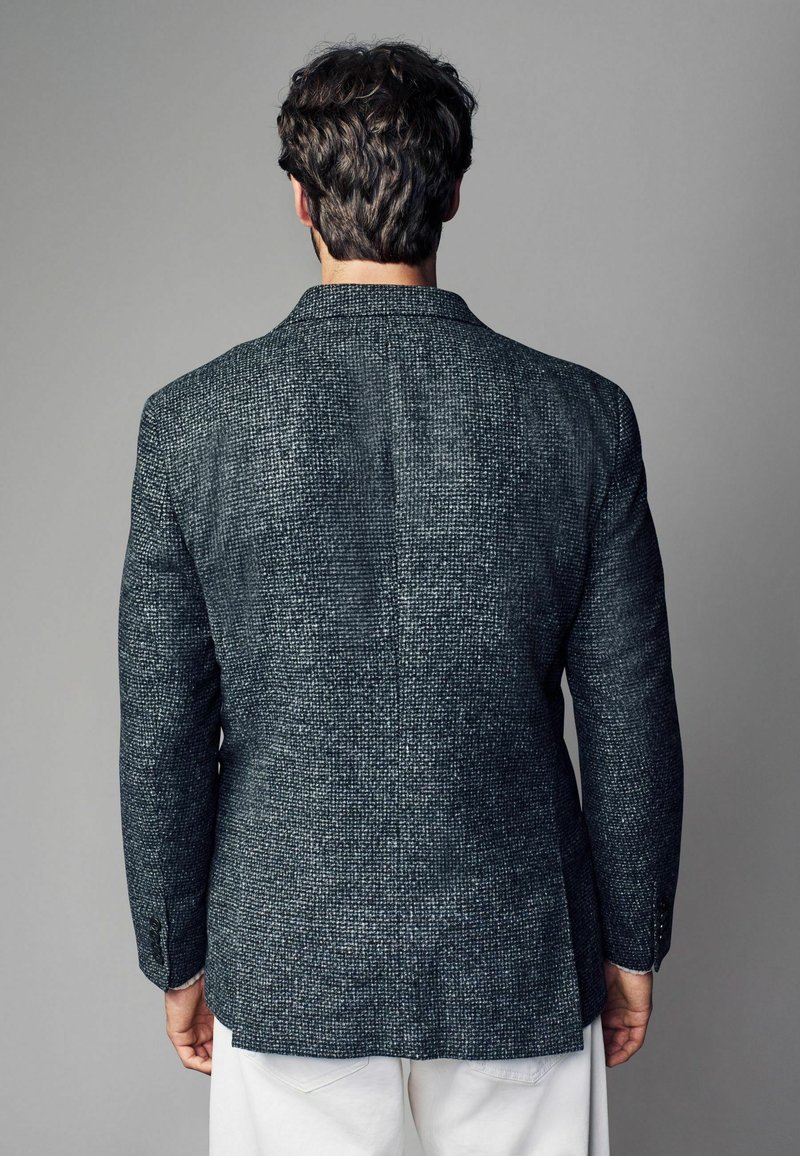 Mørk struktureret blazer med et diskret mønster, enkelt slids og spidse klapper. Stoffet ser vævet ud i en blandet grå og blå nuance.
