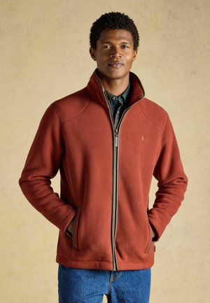 Mann trägt eine rostfarbene Fleecejacke mit Reißverschluss über einem dunkelgrünen Hemd, Hände in den Taschen, vor neutralem Hintergrund.