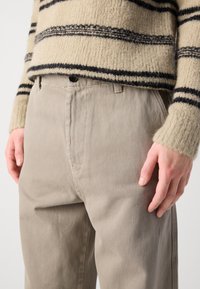 Beige puuvillahousut suoresta leikkauksesta, jotka on varustettu napituksella ja hienovaraisella tekstuurilla; yhdistetty raidoitettuun, karvaiseen villapaitaan, jonka värit ovat kerma ja musta.