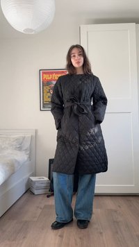 Tânără purtând o haină lungă matlasată neagră legată în talie, blugi largi albaștri și pantofi negri, stând într-un dormitor.