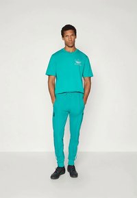 Chándal turquesa con una camiseta de manga corta y pantalones ceñidos, que presenta un logo bordado y franjas laterales; lleva zapatos negros.