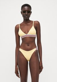 Gul bikini med triangulär topp och tunna band, prydd med en horisontell flerfärgad rand. De matchande underdelarna har en högskuren design.