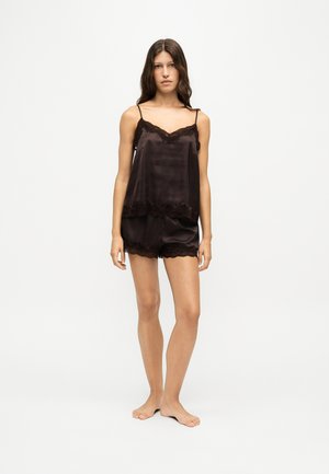 Jeune femme debout pieds nus portant un caraco en satin noir et un short assorti avec bordure en dentelle sur un fond blanc uni.